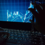Close-up of hands on a laptop keyboard while a blue stock chart with candlesticks and trend lines appears on the screen, a pen held above the keyboard.