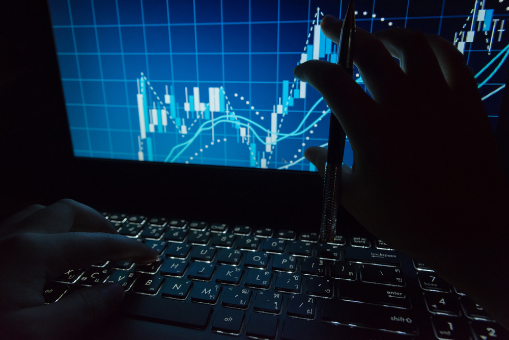 Close-up of hands on a laptop keyboard while a blue stock chart with candlesticks and trend lines appears on the screen, a pen held above the keyboard.